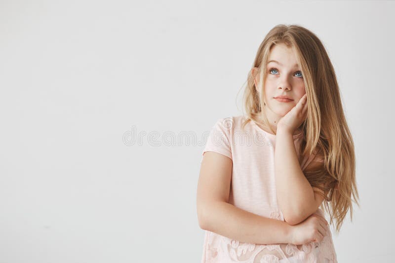 Close up of good-looking girl with blonde hair and blue eyes holding hand on cheek, looking aside with dreamy expression