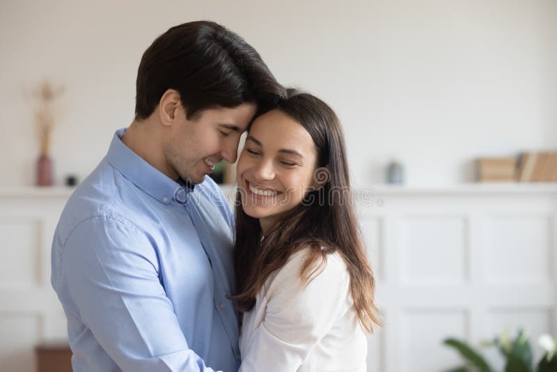 Close up happy young couple hugging, smiling women and men enjoying tender moment, standing at home, happy attractive wife and husband touching foreheads, expressing love and care, romantic date. Close up happy young couple hugging, smiling women and men enjoying tender moment, standing at home, happy attractive wife and husband touching foreheads, expressing love and care, romantic date