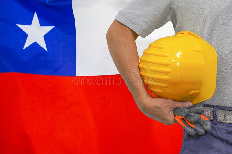 Close-up of hard hat holding by construction worker on Chile flag background. Hand of worker with yellow hard hat and gloves. Concept of Industry, construction and industrial workers in Chile. Close-up of hard hat holding by construction worker on Chile flag background. Hand of worker with yellow hard hat and gloves. Concept of Industry, construction and industrial workers in Chile