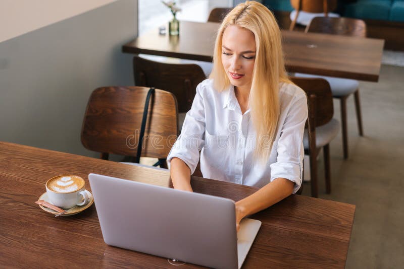 Close-up high angle view of focused blonde young woman typing something on laptop keyboard sitting at desk in coffee shop. Attractive lady chatting on laptop using dating website for acquaintance.