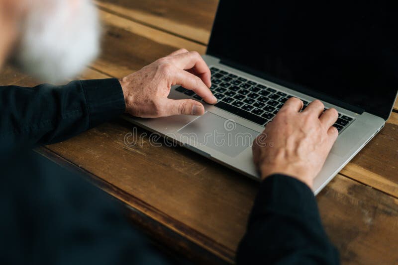 Close-up high-angle view of unrecognizable bearded mature adult male typing on laptop keyboard sitting at table at home office. Closeup of aged business man working on computer indoors, surfing online