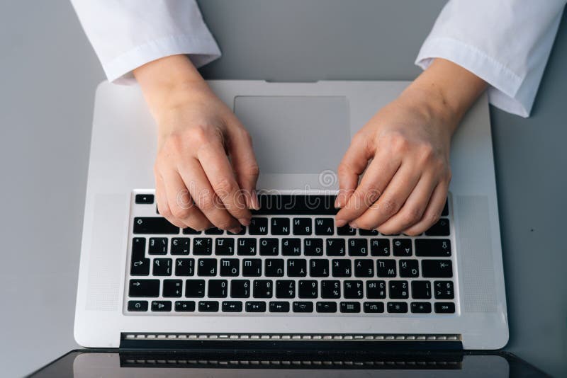 Close-up high-angle view of unrecognizable female doctor wearing white medical uniform using typing on laptop keyboard sitting at desk in modern office of medic clinic, cropped shot. top view.