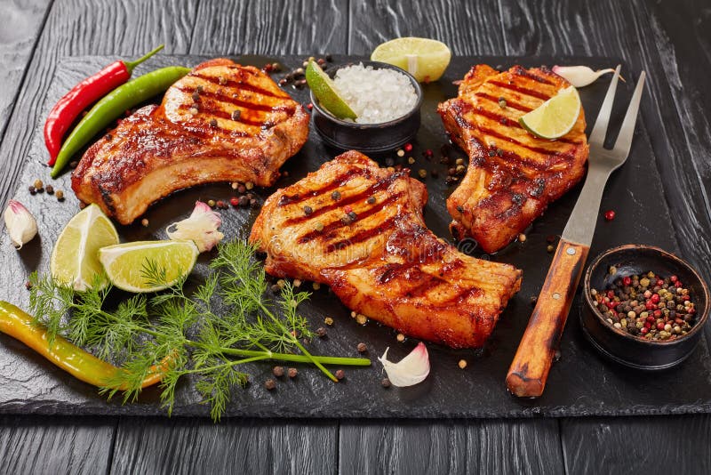 Close-up of hot grilled juicy pork chops on rib bones with spices, chili pepper, salt, lime slices and spices on black slate plate, on wooden table, view from above. Pork view stock images, royalty-free photos and pictures