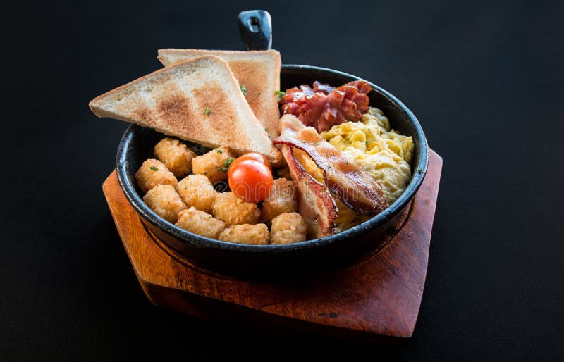 Close up image of American brunch breakfast menu with toast, tomatoes, hash brown, bacon, eggs, sausages on a black pan and wooden