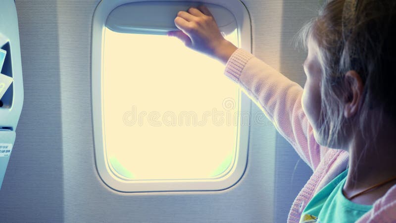 Close-up. Kid girl raises the porthole curtain in the airplane`s cabin, from there shines a bright light. girl looking