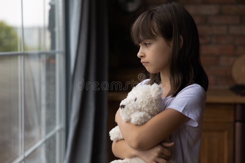 Close up lonely little girl hugging toy, looking out window