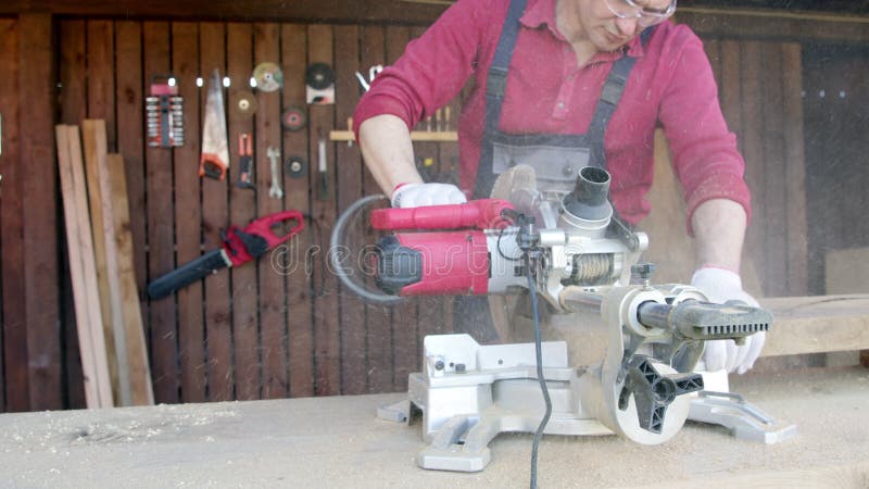 Close-up of male carpenter saws piece of beam with miter saw and flies sawdust