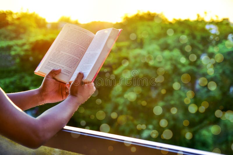 Close-up of man`s hands holding a book by the window for reading against sunset with beautiful nature background.