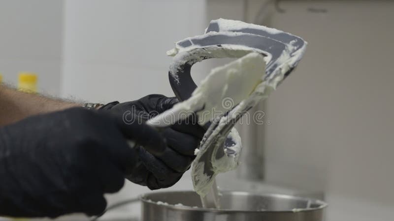 Close up of a pastry chef's gloved hands scraping dough or frosting from a mixer paddle with a spatula, emphasizing the process of preparing ingredients in a professional kitchen. Frosting spatula illustrations