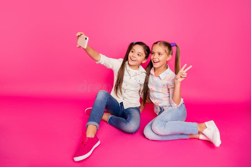 Close up photo two people little age she her girls hold hands arms telephones make take selfies v-sign sit floor wear.