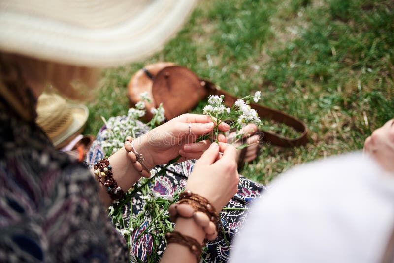 Close-up picture of woman, wearing colorful bracelets, making flower camomile wreath on green field on sunny summer day. Eco