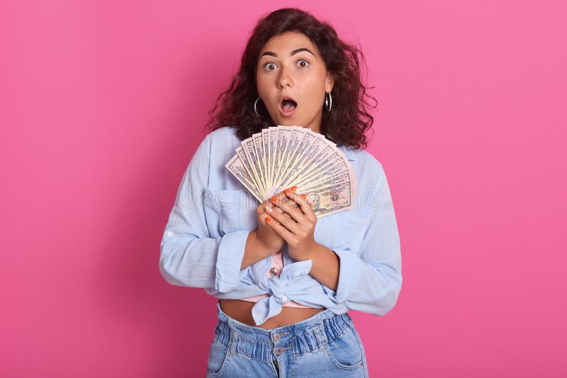 Close up portrait of attractive woman holding fan of money in her hands, keeps mouth opened, wearing stylish clothng, wins lottery, has shocked facial expression, wide open eyes with surprise