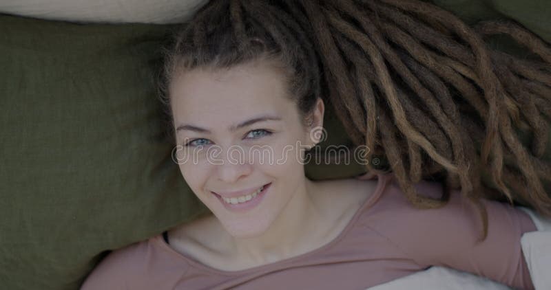 Close-up portrait of attractive young lady with dreadlocks pulling down blanket from face and smiling lying on pillow in stock video