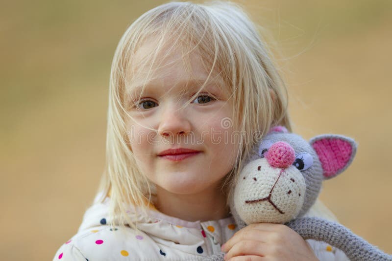 Close-up portrait of beautiful blond toddler girl hugging her favorite handmade toy knitted cat outdoors.