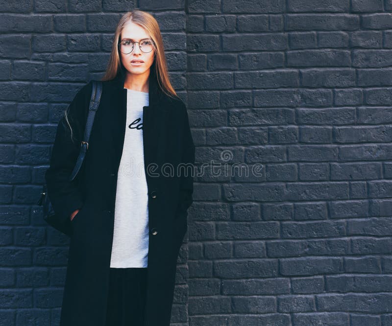Close-up portrait of a beautiful blonde girl in round fashionable glasses in a black coat and boots near a black brick royalty free stock images
