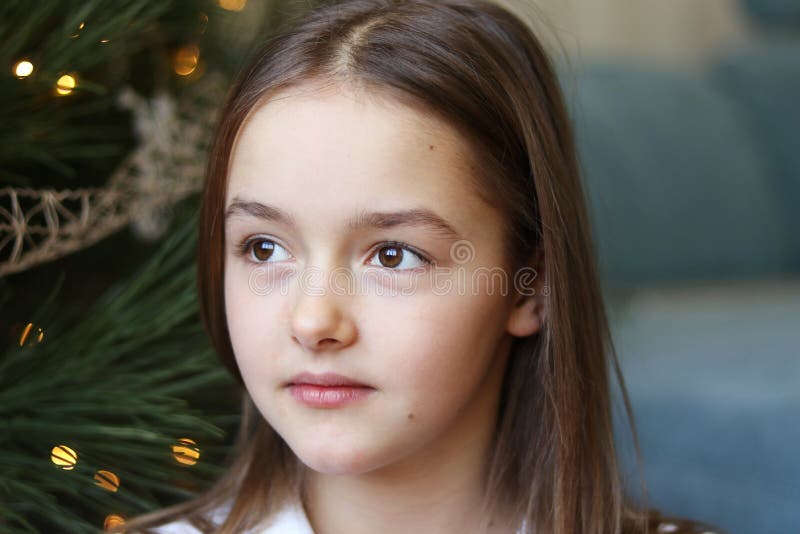 Close up portrait of beautiful little girl with brown eyes sitting under Christmas tree daydreaming