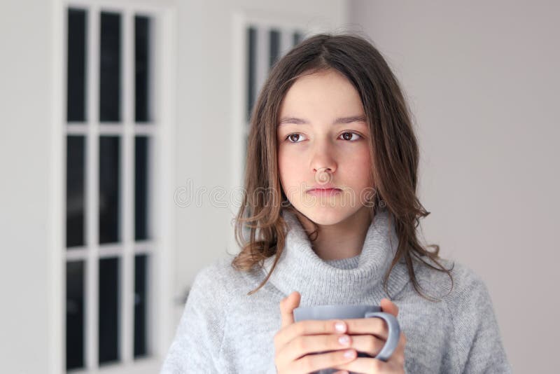 Close-up portrait of beautiful pensive tween girl in warm grey pullover holding cup of tea