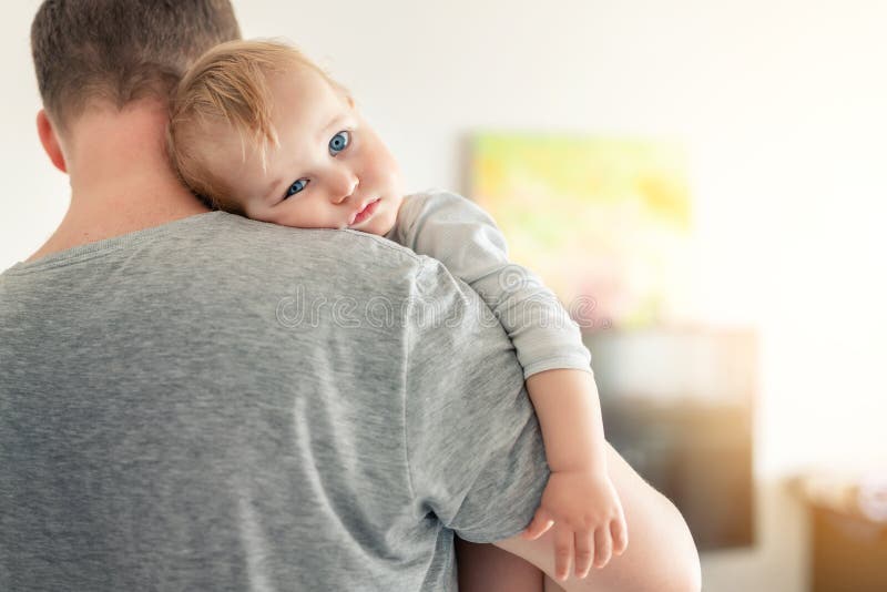 Close-up portrait of cute adorable blond caucasian toddler boy on fathers shoulder indoors. Sweet little child feeling safety on