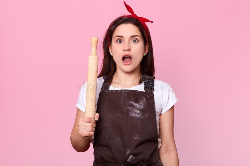 Close up portrait of emotional brunette housewife opens her mouth widely with shock, holding wooden rolling pin, astonished female royalty free stock photography Close up portrait of emotional brunette housewife opens her mouth widely with shock, holding wooden rolling pin, astonished female royalty free stock photography