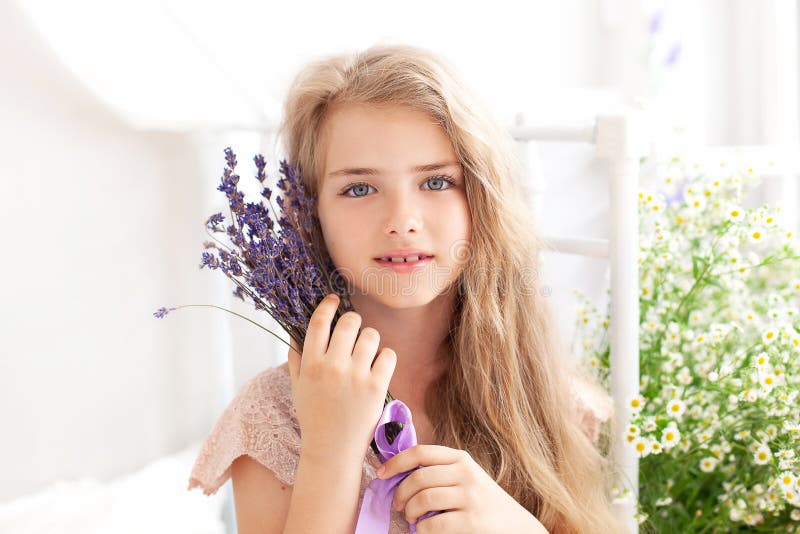 A close-up portrait of the face of a little blonde girl with long hair holding a bouquet of lavender against the background of a b