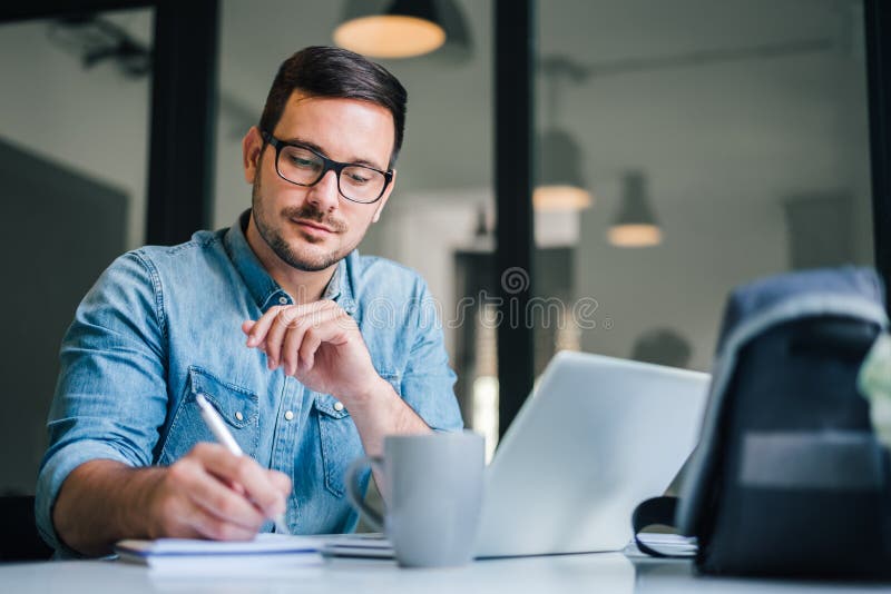 Close up portrait of handsome man working from home office taking reading and writing notes in note pad while working on laptop