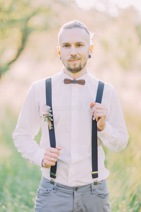 The close-up portrait of the modern-dressed groom in the gray pants, white shirt and wooden butterfly at the background stock photography The close-up portrait of the modern-dressed groom in the gray pants, white shirt and wooden butterfly at the background stock photography