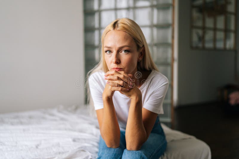 Close-up portrait of pensive young blonde woman sitting alone in living room and sad looking at camera holding hands on