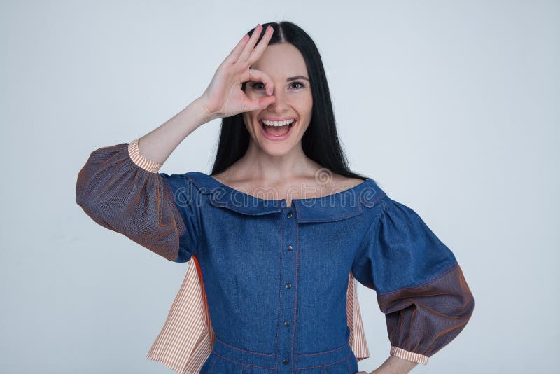 Close up portrait of smiling brunette woman with white teeth, looking at the camera through fingers in ok gesture. wearing fashion stock photos Close up portrait of smiling brunette woman with white teeth, looking at the camera through fingers in ok gesture. wearing fashion stock photos