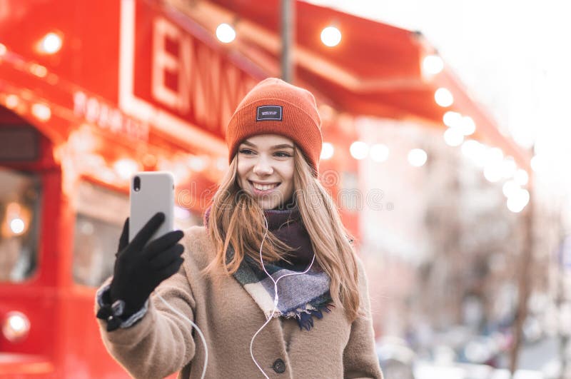 Close-up portrait of a smiling girl in warm clothes takes selfie on a smartphone with a bright red city background. Beautiful girl royalty free stock photo Close-up portrait of a smiling girl in warm clothes takes selfie on a smartphone with a bright red city background. Beautiful girl royalty free stock photo