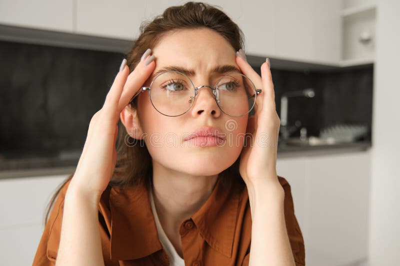 Close up portrait of woman in glasses, holding hands on head and frowning from painful headache, having problem, thinking about concerning situation.