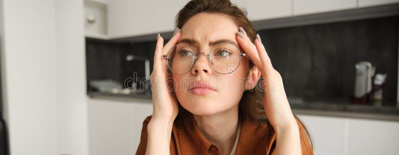 Close up portrait of woman in glasses, holding hands on head and frowning from painful headache, having problem, thinking about concerning situation.
