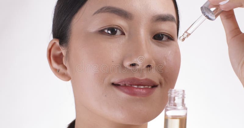 Close up portrait of young asian woman applying serum on her cheek, skin care, white studio background