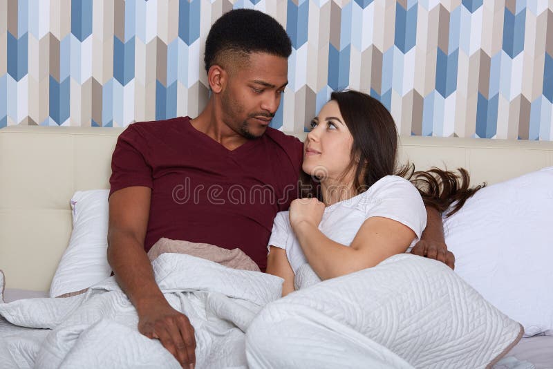 Close up portrait of young male and female lying in white bed togethe, wearing pajamas. Romantic couple in love looking at each