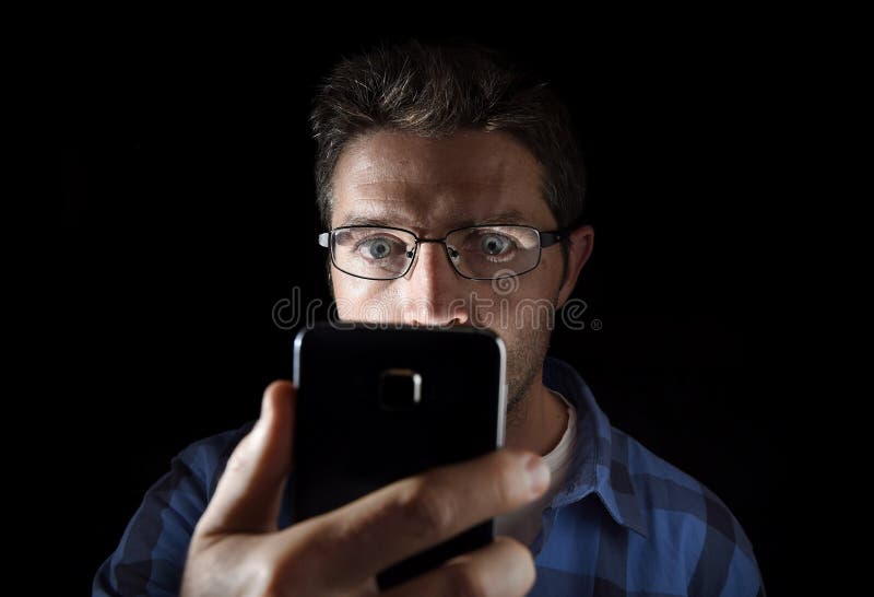 Close up portrait of young man looking intensively to mobile phone screen with blue eyes wide open isolated on black background
