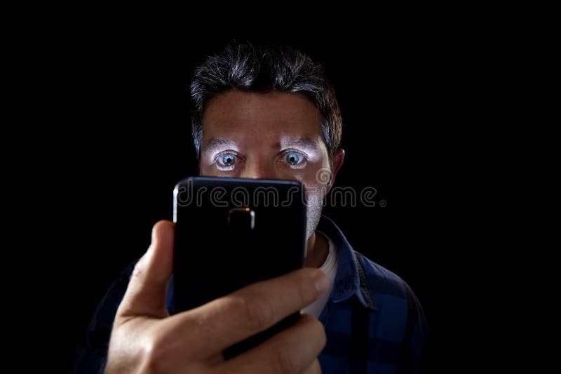 Close up portrait of young man looking intensively to mobile phone screen with blue eyes wide open isolated on black background