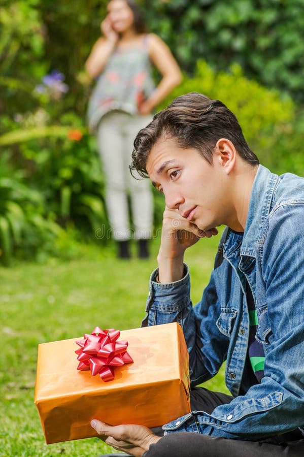 Close up of sad man wearing a jean jacket and black pants sitting in the ground holding gift with a blurred woman behind royalty free stock photography Close up of sad man wearing a jean jacket and black pants sitting in the ground holding gift with a blurred woman behind royalty free stock photography