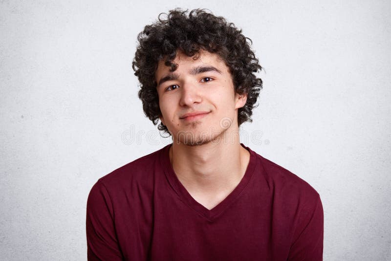 Close up shot of curly haired European teenager with little beard, wears maroon shirt. Young smiling male stands with lowered