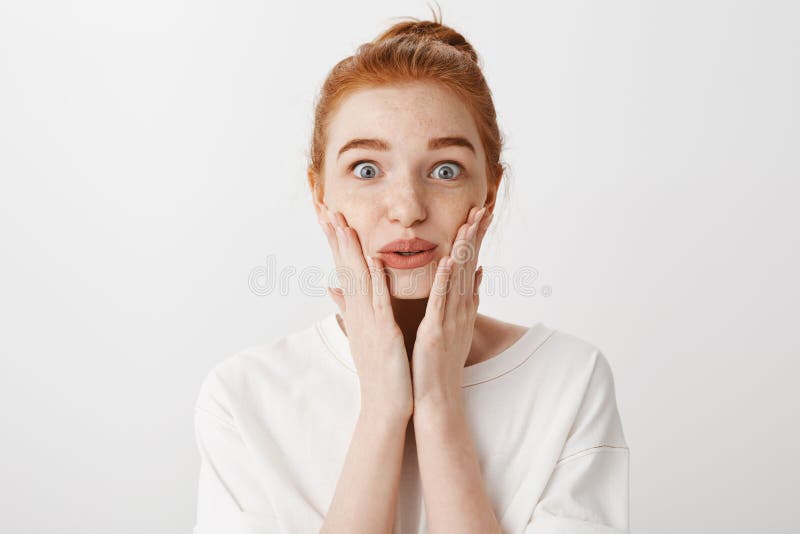 Close-up shot of impressed sympathizing redhead woman with bun hairstyle, squeezing lips while holding palms on cheeks, being shocked and stunned with terrifying news on tv, standing over grey wall.