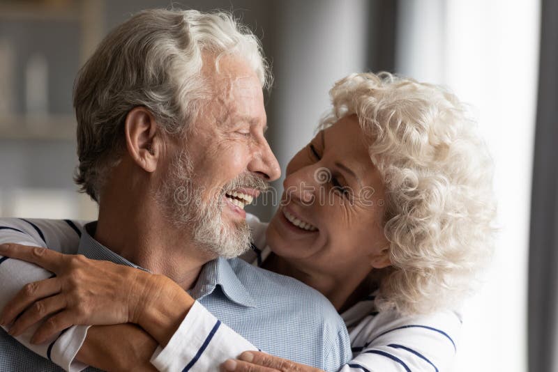 Close up smiling older couple hugging, happy senior family enjoying tender moment together, loving caring mature wife embracing husband from back, looking at each other, eternal love. Close up smiling older couple hugging, happy senior family enjoying tender moment together, loving caring mature wife embracing husband from back, looking at each other, eternal love