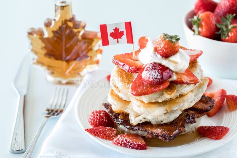 A close up of a stack of pancakes with a bottle of syrup and bowl of strawberries in behind. Canada Day concept.