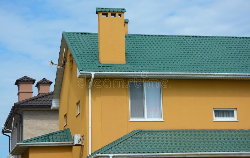 A close-up on a stucco, plastered and painted house of ochre color with green metal roof, chimney and white rain gutters with a