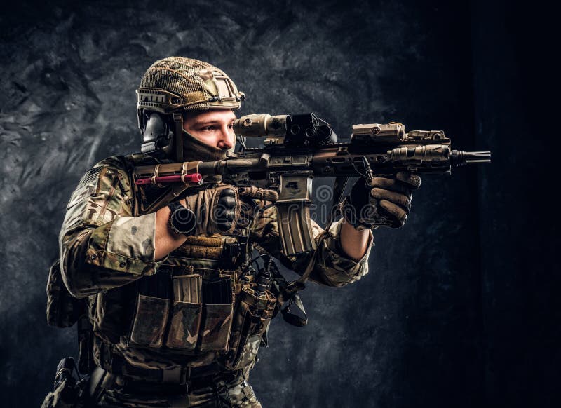 Close-up studio photo against a dark wall. The elite unit, special forces soldier in camouflage uniform holding an royalty free stock photo Close-up studio photo against a dark wall. The elite unit, special forces soldier in camouflage uniform holding an royalty free stock photo