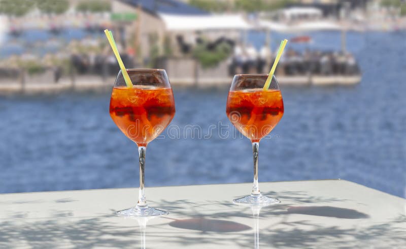 Close up of two glasses of chilled cocktail drinks with straws and ice cubes on a table near the beach.