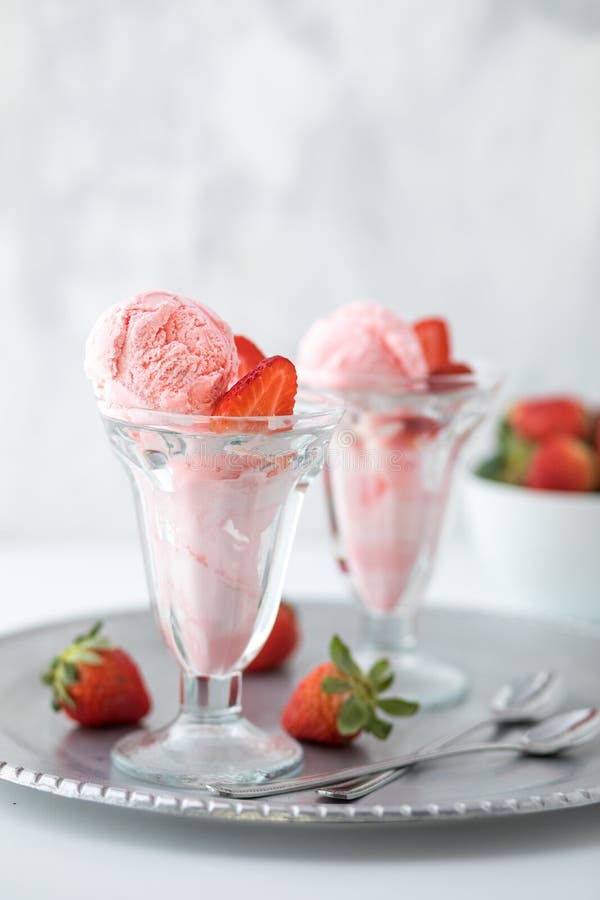 Two strawberry ice cream sundaes on a metal tray with a bowl of strawberries in behind.