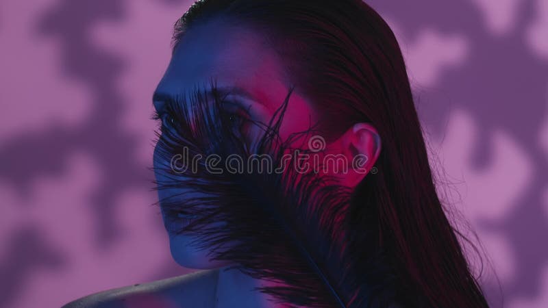 Close up video of a young woman calmly looking away, half a turn portrait, feather moving by the face, neon solor scheme