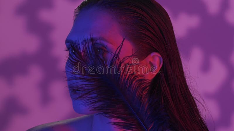 Close up video of a young woman calmly looking away, half a turn portrait, feather moving by the face, neon solor scheme