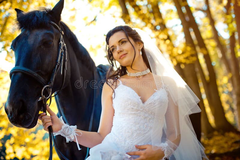 Close-up wedding portrait of the pretty lovely brunette bride tenderly smiling and posing with the horse in the autumn