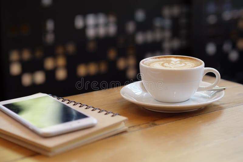 Close up white coffee cup with latte art on wood table with smartphone at coffee shop