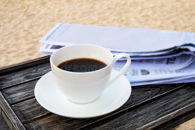 Close up white coffee cup on wood table at sunrise sand beach with newspaper in the morning