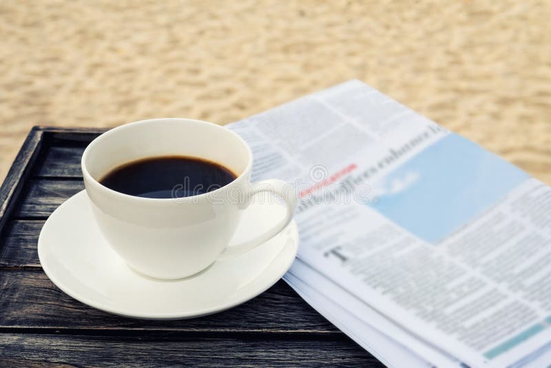 Close up white coffee cup on wood table at sunrise sand beach with newspaper in the morning, warm tone